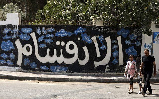 رجل فلسطيني وطفلتيه يمران من أمام رسم غرافيتي كُتب عليه بالعربية ’الإنقسام’ في مدينة غزة، في 17 سبتمبر، 2017، بعد أن أعلنت حركة ’حماس’ عن موافقتها على خطوات للسماح بحل النزاع مع حركة ’فتح’ المستمر منذ عقد من الزمن واستعدادها لإجراء انتخابات. (AFP/Mahmud Hams) 