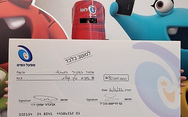 رجل حصل على 80 مليون شيكل في أكبر جائزة لوتو إسرائيلية يقف مع شيك ضخم في مكاتب مفعال هبايس في 6 فبراير، 2022. (Mifal HaPais)