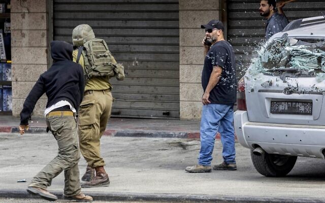 مستوطن إسرائيلي يلقي حجرا لكسر الزجاج الخلفي لسيارة فلسطينية أثناء وقوف جندي إسرائيلي بجانبه، خلال مواجهات في بلدة حوارة بالضفة الغربية، 13 أكتوبر 2022 (Oren ZIV / AFP)