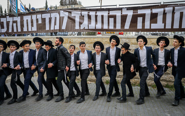 رجال وشباب من الحريديم يرقصون أمام لافتة كتب عليها "يجب أن نقاوم دولة الشريعة"، بينما يحتشد المتظاهرون خارج منزل زعيم حزب "شاس" أرييه درعي في القدس، 23 مارس 2023 (Flash90)