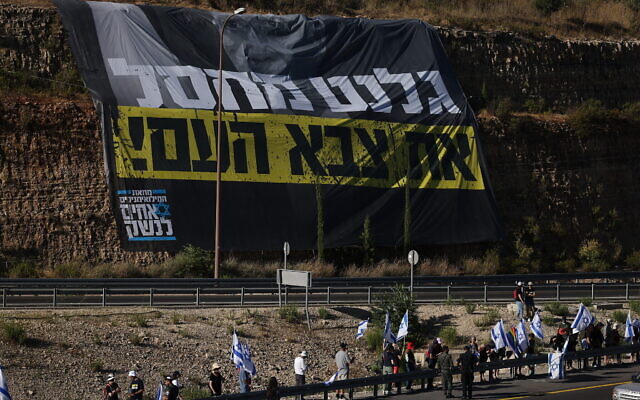 متظاهرون يلوحون بالأعلام الإسرائيلية أثناء احتجاجهم على خطط الحكومة لإصلاح النظام القضائي، عند الطريق رقم 1 بالقرب من عين حميد، خارج القدس، 18 يوليو 2023 (Yonatan Sindel / Flash90)