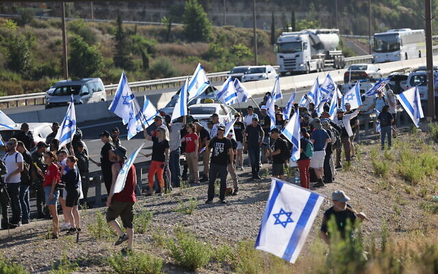 متظاهرون يلوحون بالأعلام الإسرائيلية أثناء احتجاجهم على خطط الحكومة للإصلاح القضائي ، عند الطريق رقم 1 بالقرب من عين حميد ، خارج القدس ، 18 يوليو ، 2023 (Yonatan Sindel / Flash90)