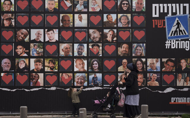 امرأة وأطفالها يسيرون أمام جدار عليه صور الرهائن الذين تم اختطافهم خلال الهجوم عبر الحدود الذي قادته حماس في إسرائيل في 7 أكتوبر، 26 فبراير، 2024. (Leo Correa/AP)