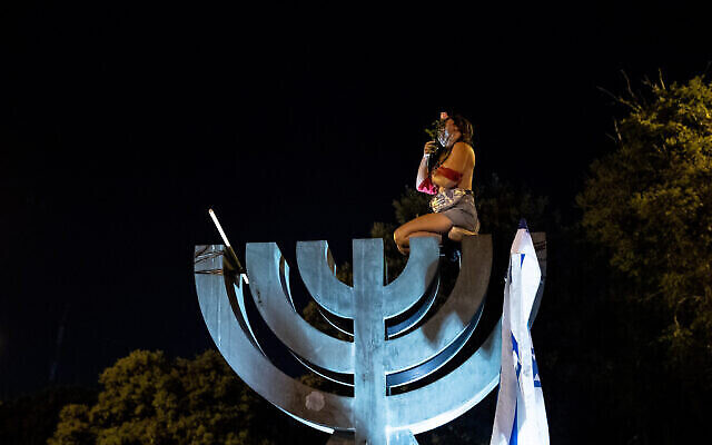 A topless protester sits on a menorah statue near the Knesset during a demonstration against Prime Minister Benjamin Netanyahu in Jerusalem on July 21, 2020. (Yonatan Sindel/Flash90)