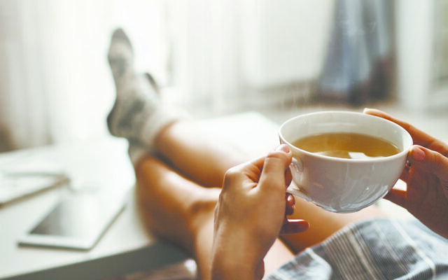 Cup of tea and chill. Woman lying on couch, holding legs on coffee table, drinking hot coffee and enjoying morning, being in dreamy and relaxed mood. Girl in oversized shirt takes break at home.