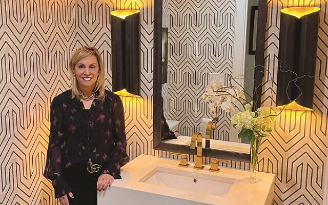 Andrea Turry poses in a powder room she designed using hexagon floor tiles, a graphic wall covering, forged brass vanity with a quartz top, oversized sconces and a custom designed mirror.