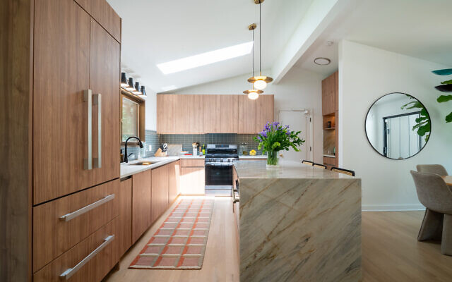 The Mid Century Modern  kitchen has warm wooden cabinetry and a waterfall quartzite island in distinctive earthy color tones. Laura’s arsenal: a secret spice drawer is situated next to the stove.  