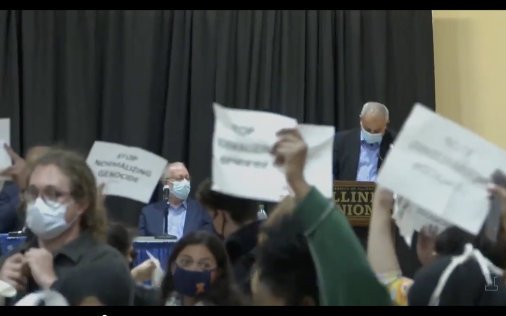 Students at University of Illinois Urbana-Champaign holding signs that read 'STOP NORMALIZING GENOCIDE' stage a walk-out at the event 'Remaining In Dialogue,' with Yossi Klein Halevi (seated) and Mohammad Darawshe (at lecturn), October 7, 2021 (Screen grab from a video of the event)