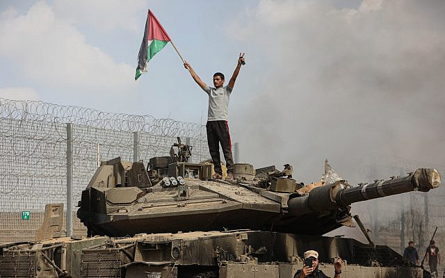 A Palestinian stands on a burning tank after some 3,000 Hamas terrorists destroyed the border fence and entered Israel. October 7, 2023. (Yousef Mohammed/Flash90) - Source: The Times of Israel.
