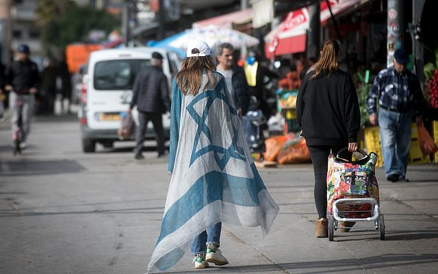 A woman wearing a large Israeli flag walks through the Carmel market in Tel Aviv. December 31, 2023. (Miriam Alster/FLASH90)