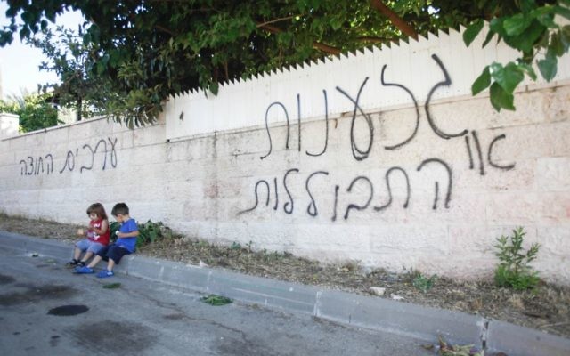 Des enfants assis près d'un mur où il est inscrit en hébreu : "racisme ou assimilation", "les Arabes dehors" à Abu Ghosh (Crédit : Flash90)