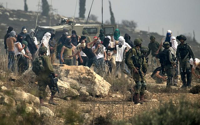 Photo d'illustration : Des soldats israéliens regardent des habitants d'implantation masqués jeter des pierres à des manifestants palestiniens (non-visibles sur la photo) pendant une manifestation contre la construction d'un avant-poste israélien aux abords du village de Turmusaya et de l'implantation de Shilo, au nord de Ramallah, en Cisjordanie, le 17 octobre 2019. (Crédit : JAAFAR ASHTIYEH / AFP)