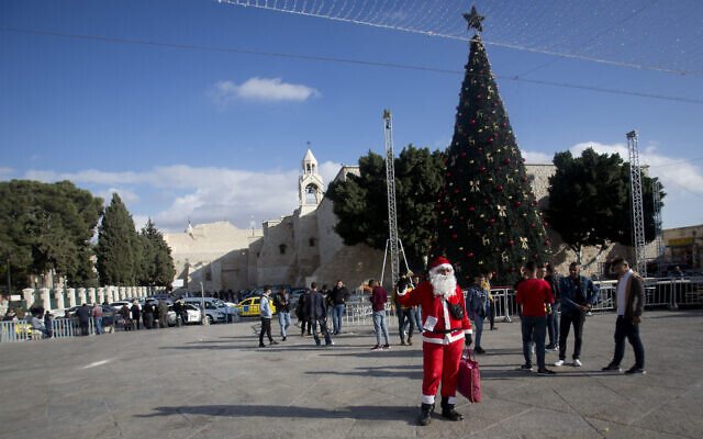 Un Palestinien portant un costume de père Noël accueille des visiteurs chrétiens à l'extérieur de l'église de la Nativité, traditionnellement considérée par les Chrétiens comme le lieu de naissance de Jésus-Christ, dans la ville cisjordanienne de Bethléem, le 5 décembre 2019. (Crédit : Majdi Mohammed / AP)