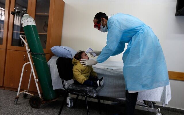 Un agent de santé palestinien portant un masque de protection vérifie la température corporelle d'un enfant dans une école de l'UNRWA dans le camp de réfugiés d'al-Shati à Gaza, le 18 mars 2020, en pleine pandémie de coronavirus. (Crédit : MAHMUD HAMS / AFP)