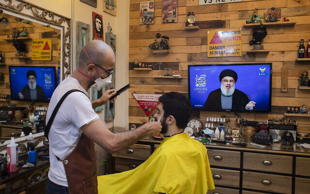 Un barbier portant un masque pour limiter la propagation du nouveau coronavirus écoute un discours du chef du Hezbollah Sayyed Hassan Nasrallah diffusé sur la chaîne de TV du Hezbollah  al-Manar, pour marquer la Journée Al-Quds (Jérusalem) alors qu'il coupe les cheveux d'un client dans son salon situé dans la banlieue sud de Beyrouth au Liban, le vendredi 22 mai 2020. (AP Photo/Hassan Ammar)