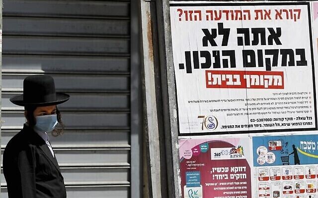 Un ultra-orthodoxe portant un masque passe devant une pancarte disant : "Vous lisez cette pub ? Vous n'êtes pas au bon endroit. Le bon endroit, c'est chez vous", dans la ville religieuse de Bnei Brak, près de Tel Aviv, le 3 avril 2020. (Crédit : JACK GUEZ / AFP)