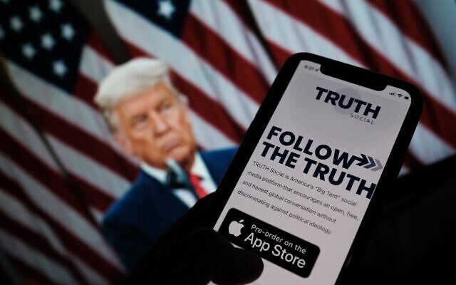 Illustration : une personne consultant une boutique d'applications sur un smartphone pour "Truth Social", avec une photo de l'ancien président américain Donald Trump sur un écran d'ordinateur en arrière-plan, à Los Angeles, le 20 octobre 2021. (Crédit : Chris DELMAS / AFP)