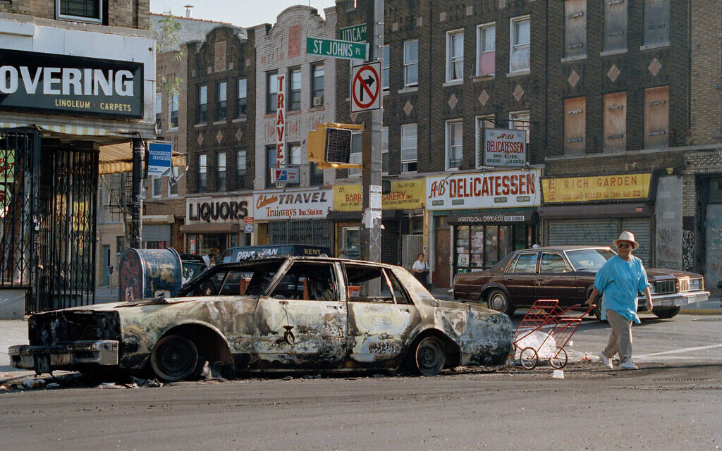 Une femme marchant devant une voiture incendiée en traversant St. Johns Place sur Utica Avenue dans le quartier de Crown Heights, dans l'arrondissement de Brooklyn à New York le 22 août 1991. Photo d'archive (Crédit : David Cantor/AP Photo)