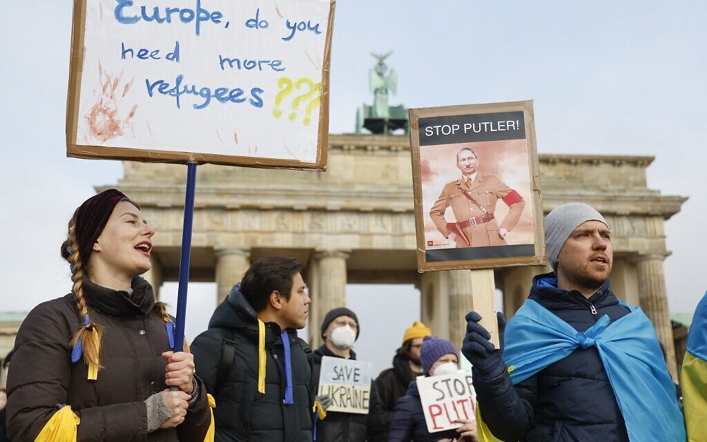 Des manifestants enveloppés dans des drapeaux ukrainiens tiennent des pancartes sur lesquelles on peut lire "Stop Putler" et "Europe, avez-vous besoin de plus de réfugiés ?" devant la Porte de Brandebourg à Berlin pour manifester en faveur de la paix en Ukraine, le 27 février 2022. (Crédit : Odd ANDERSEN / AFP)