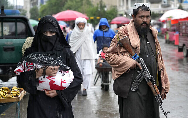 Une Afghane marche dans le vieux marché, alors qu’un combattant taliban monte la garde, au centre-ville de Kaboul, en Afghanistan, le 3 mai 2022. (Crédit : AP Photo/Ebrahim Noroozi)