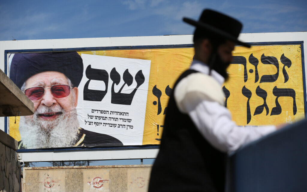 Illustration : Un Juif ultra-orthodoxe marchant près d'une photo du défunt chef spirituel du Shas, le rabbin Ovadia Yosef, sur un panneau d'affichage dans le cadre de la campagne électorale de Shas, dans la ville de Safed, au nord d'Israël, le 28 février 2020. (Crédit : David Cohen/Flash90)