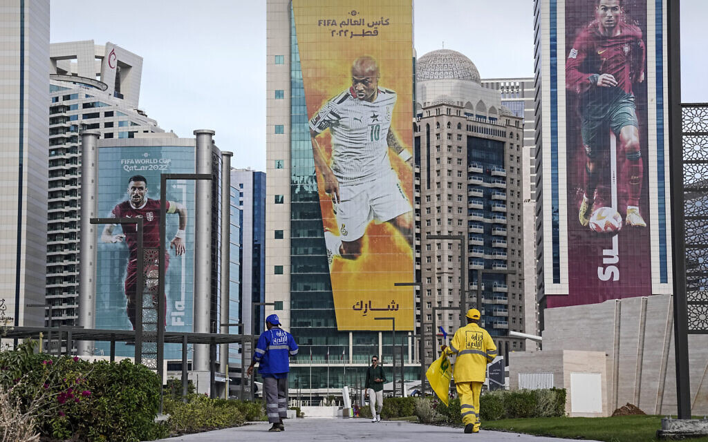 Deux ouvriers nettoient une rue devant des gratte-ciel avec des affiches géantes des stars du football Cristiano Ronaldo du Portugal, Andre Ayew du Ghana et Dusan Tadic de Serbie, à Doha, au Qatar, le 29 novembre 2022. (Crédit : AP/Martin Meissner)