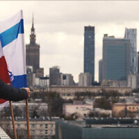 Jonny Daniels se tenant à côté des drapeaux israélien et polonais sur le toit d'un immeuble résidentiel surplombant ce qui fut le ghetto de Varsovie, à Varsovie, en Pologne, le 19 mars 2023. (Crédit : From the Depths)