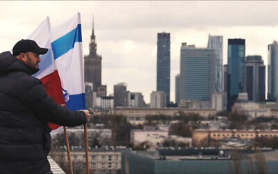 Jonny Daniels se tenant à côté des drapeaux israélien et polonais sur le toit d'un immeuble résidentiel surplombant ce qui fut le ghetto de Varsovie, à Varsovie, en Pologne, le 19 mars 2023. (Crédit : From the Depths)