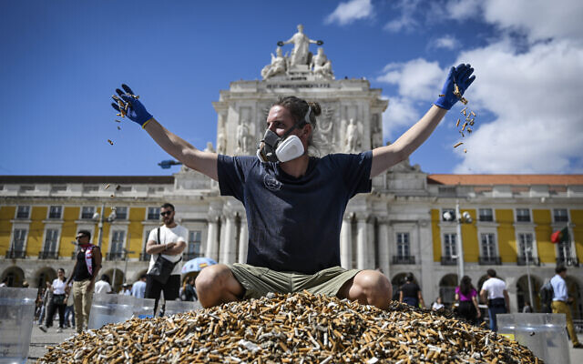 Le militant écologiste allemand, Andreas Noe, 34 ans, jette des mégots de cigarettes alors qu'il est assis au-dessus d'un tas de mégots, ramassés en une semaine, sur la place Comercio à Lisbonne le 23 avril 2023. (Crédit : PATRICIA DE MELO MOREIRA / AFP)