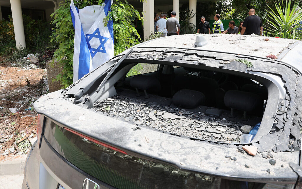 Un drapeau national israélien suspendu derrière une voiture endommagée, devant un bâtiment dans lequel une personne a été tuée après avoir été touchée la veille par une roquette tirée depuis la Bande de Gaza, à Rehovot, le 12 mai 2023. (Crédit : Jack Guez/AFP)