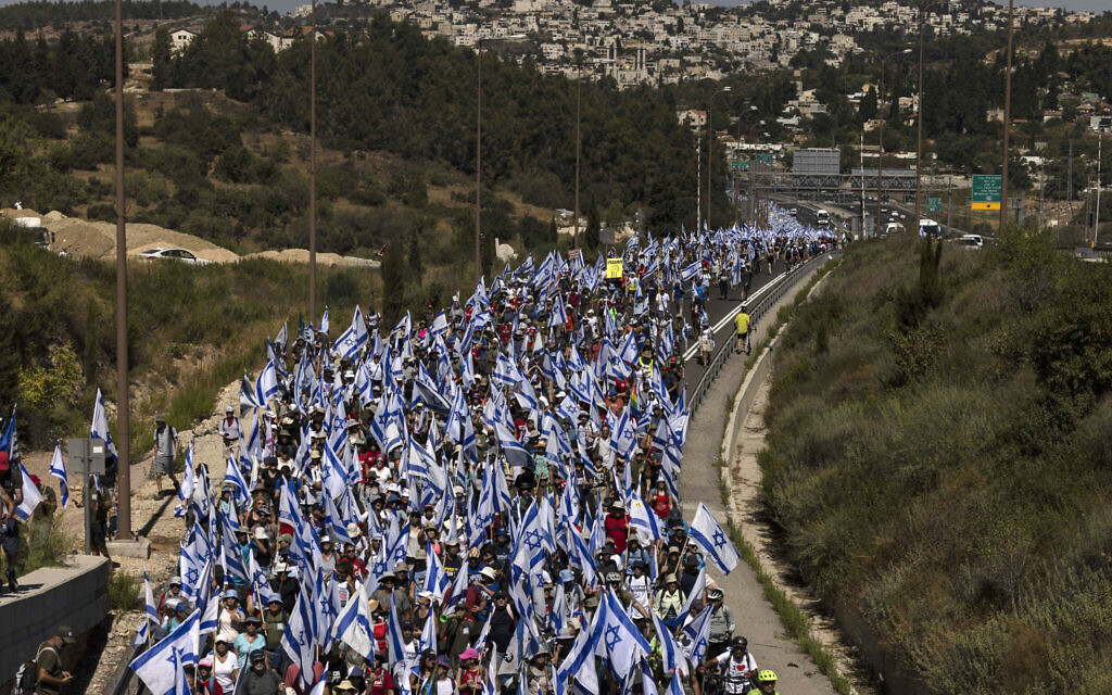  Des manifestants brandissant des drapeaux nationaux israéliens, lors d'une marche de plusieurs jours de Tel Aviv à Jérusalem pour protester contre le projet de loi du gouvernement sur la réforme judiciaire avant un vote au Parlement alors qu'ils marchent sur l'autoroute près de la ville de Mevasseret Zion, le 22 juillet 2023. (Crédit : Menahem Kahana/AFP)