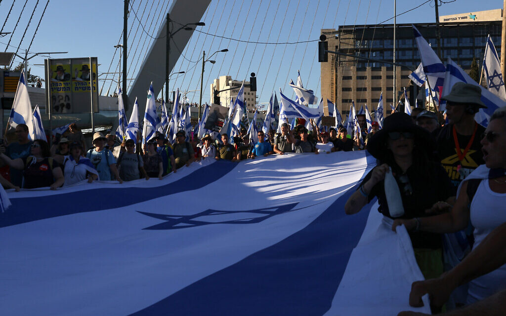  Des manifestants portant un grand drapeau israélien alors qu'ils marchent vers Jérusalem, au cours d'une marche de plusieurs jours qui a commencé à Tel Aviv pour protester contre la législation de refonte judiciaire du gouvernement avant un vote sur la première loi de ce type à la Knesset,  le 22 juillet 2023. (Crédit :  Hazem Bader/AFP)