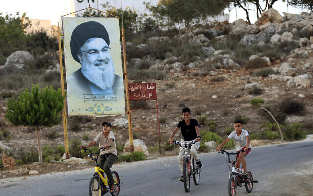 Des enfants passant à vélo devant une affiche du chef du Hezbollah, Hassan Nasrallah, dans le village frontalier libanais de Marwaheen, le 12 juillet 2023. (Crédit : Mohammed Zaatari/AP)