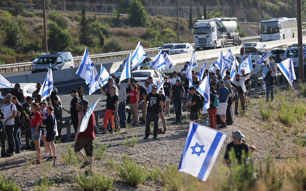 Des manifestants brandissant des drapeaux israéliens alors qu'ils protestent contre les projets de refonte judiciaire du gouvernement sur la Route 1 près d'Ein Hemed, à l'extérieur de Jérusalem, le 18 juillet 2023.. (Crédit : Yonatan Sindel/Flash90)