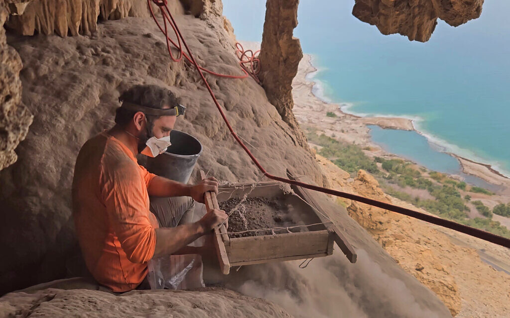 Des archéologues tamisant la terre de la grotte du désert de Judée où quatre épées romaines ont été découvertes, avec vue sur la mer Morte. (Crédit : Matan Toledano/IAA)