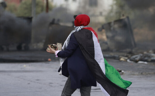 Une femme drapée dans un drapeau palestinien se préparant à lancer des pierres en direction des forces israéliennes lors d'affrontements avec elles à l'entrée nord de la ville de Ramallah, en Cisjordanie, le 20 octobre 2023 (Crédit : Jaafar Ashtiyeh/AFP)