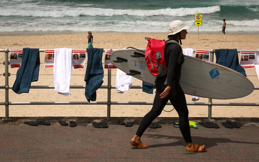 Une surfeuse avec sa planche passe devant des affiches, des serviettes et des tongs qui font partie d'une manifestation organisée par la communauté juive australienne à Bondi Beach à Sydney le 2 novembre 2023 pour souligner le sort des otages détenus par le Hamas. (Crédit : DAVID GRAY / AFP).