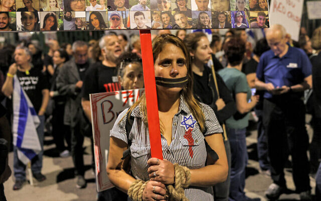 Une femme se tenant debout avec une fermeture éclair symbolique sur la  bouche et une pancarte identifiant certains des otages enlevés par des terroristes palestiniens lors de l'attaque du 7 octobre, lors d'une veillée exigeant leur retour, devant la Knesset à Jérusalem, le 7 novembre 2023. (Crédit : Ahmad Gharabli/AFP)
