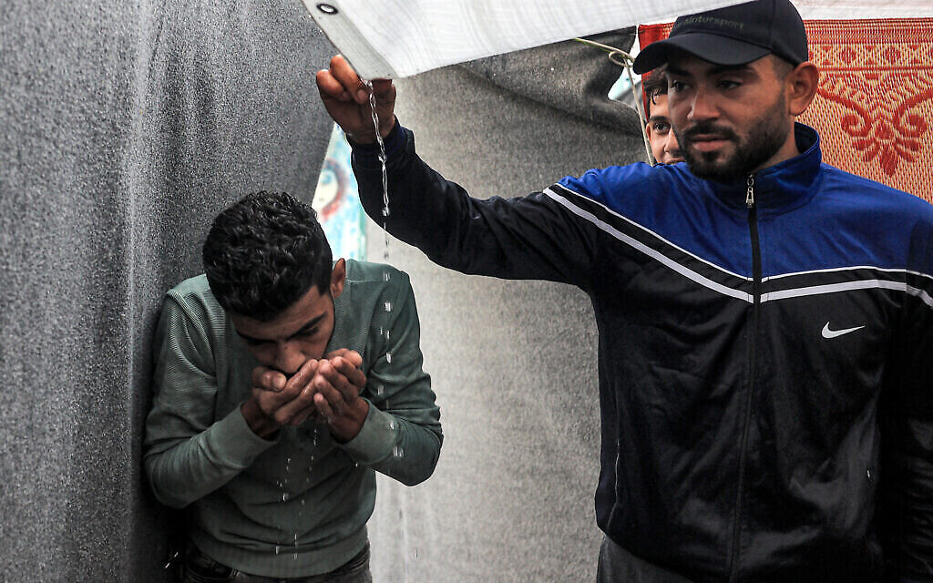 Des hommes utilisent leurs mains pour boire l'eau de pluie qui s'écoule du toit d'une tente en raison des pénuries d'eau, dans une école de l'Office de secours et de travaux des Nations Unies pour les réfugiés de Palestine dans le Proche-Orient (UNRWA) à Rafah, dans le sud de la bande de Gaza, reconvertie en abri pour les Palestiniens déplacés par la guerre entre Israël et le Hamas. Le 14 novembre 2023. (Crédit : Saïd Khatib / AFP)