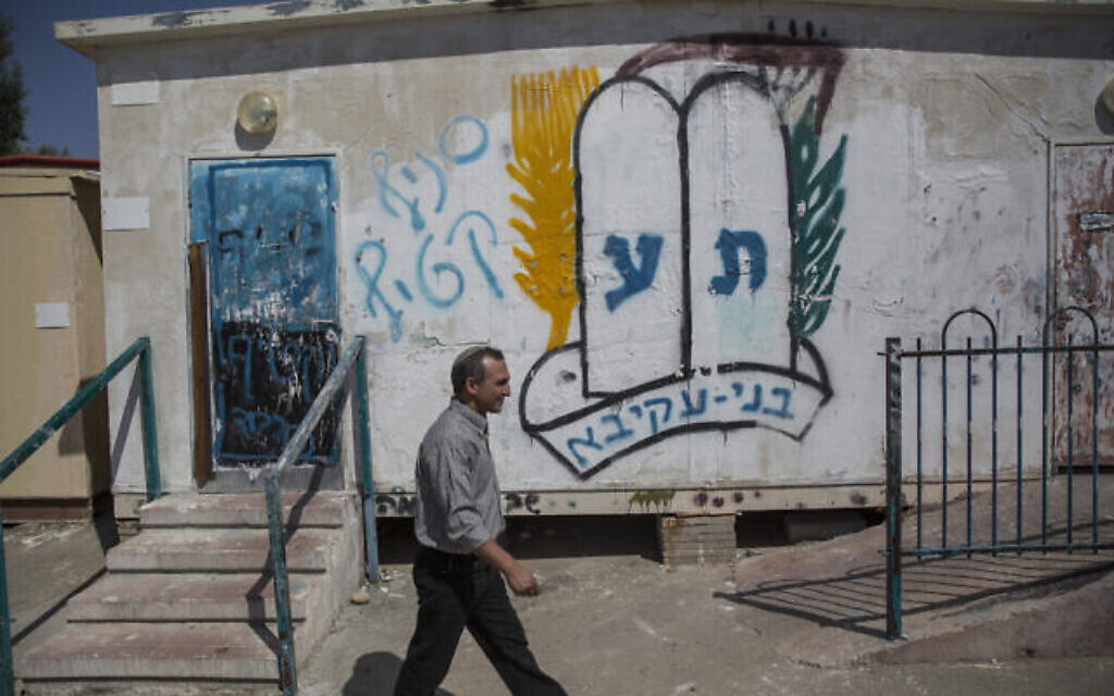 Yoram Ohev-Tzion, secrétaire de la ville de Karmei Katif, passe devant des hangars abritant le siège temporaire de la communauté Karmei Katif, près de la ville israélienne d'Amatzia, dans la région sud du mont Hébron. Sur le mur est inscrit au pochoir le logo du mouvement de jeunesse B'nei Akiva, à côté des mots « branche Katif ». 20 juillet 2015. (Crédit : Hadas Parush/Flash90)