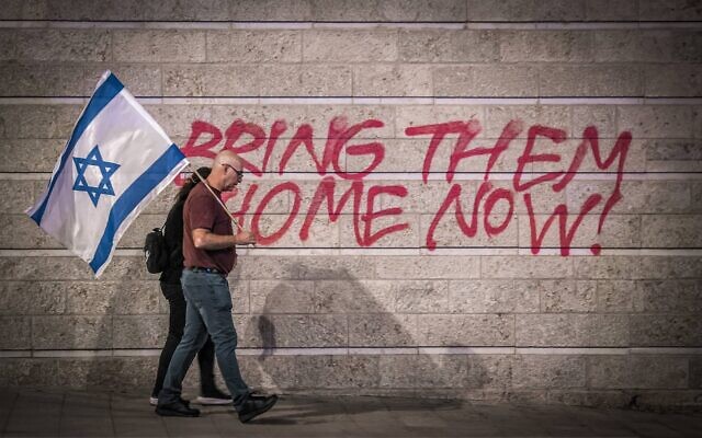Un couple d'Israéliens tenant un drapeau israélien à Jérusalem, marchant devant un graffiti appelant à la libération des otages israéliens détenus dans la bande de Gaza depuis le 7 octobre 2023 par le Hamas (Crédit : Gil Cohen-Magen / AFP)