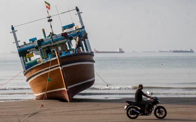 Sur cette photo de l'agence de presse iranienne ISNA, datée du 24 avril 2026, un Iranien passe à moto devant un bateau sur la plage de Suru à Bandar Abbas, le long du détroit d'Ormuz. (Razieh Poudat/ISNA/AFP)