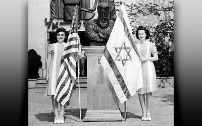 Two young women hold flags next to the podium as the Jewish-Palestine Pavilion opened, 84 years ago this month. Maurice Ascalon’s copper sculpture stands above them; a sculpture of Theodor Herzl is between them.