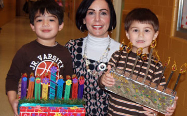 JCC preschool teacher Mindy Katz presents two of her artists, Noah Rotondo, left, with his craft stick menora, and Hayden Cear with his menora, made of twigs and pipe cleaners.