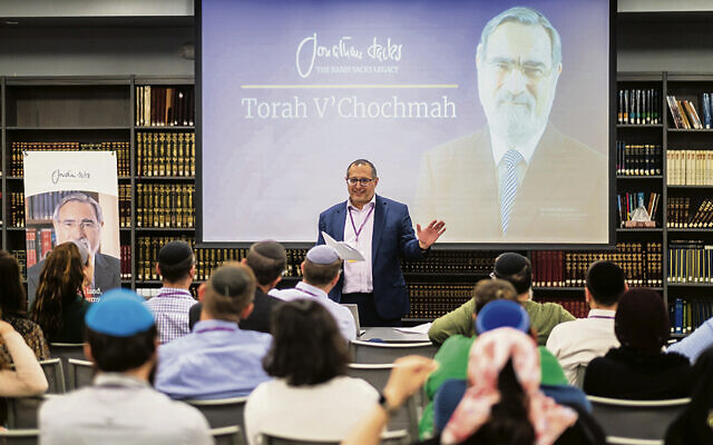 Rabbi Jeremy Bruce talks to the group; he’s standing in front of and next to photos of Rabbi Jonathan Sacks.