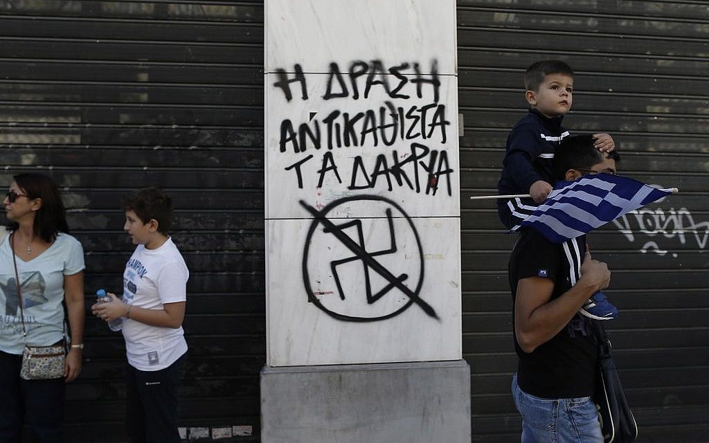 A child holds a Greek flag as he passes with his father next to graffiti depicting an anti Nazi Symbol and reading in Greek 'Action replace tears' during a parade commemorating Greece's entry in World War II in 1940, in central Athens, on Monday, Oct. 28, 2013. (photo credit: AP Photo / Petros Giannakouris)