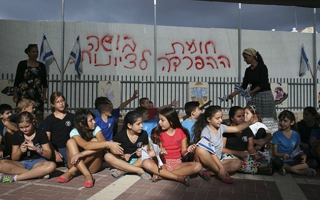 School children wave the Israeli flag as they sit in their school playground in front of a dividing barrier put up by the Beit Shemesh municipality, September 02, 2014. Graffiti  reads "Separation barrier, a disgrace to Zionism" . (photo credit: Nati Shohat/Flash90)