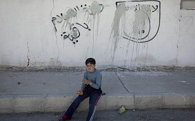 In this photo taken Oct. 29, 2014, a Jordanian boy sits on the sidewalk by painted-over graffiti depicting the flag of the Islamic State group with Arabic that reads "their is only one God and Muhammad is his prophet, the Islamic state is staying," in the city of Ma'an, Jordan,(Photo credit: AP/Nasser Nasser)