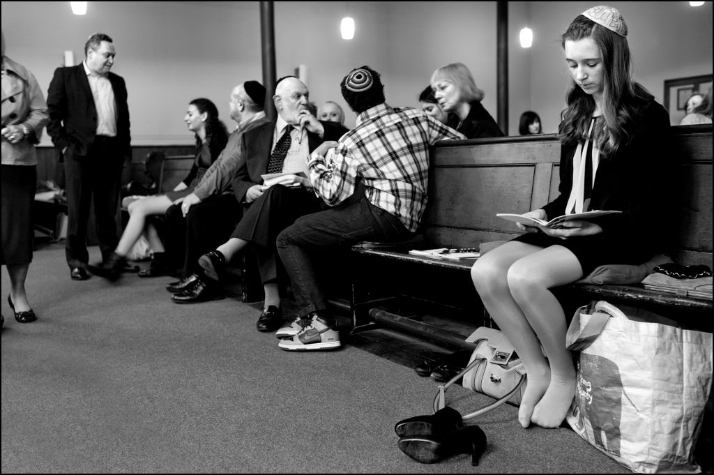 Scottish Jews at synagogue. Off to one side, a girl looks into her prayer book as other congregants talk to one another. (Judah Passow)