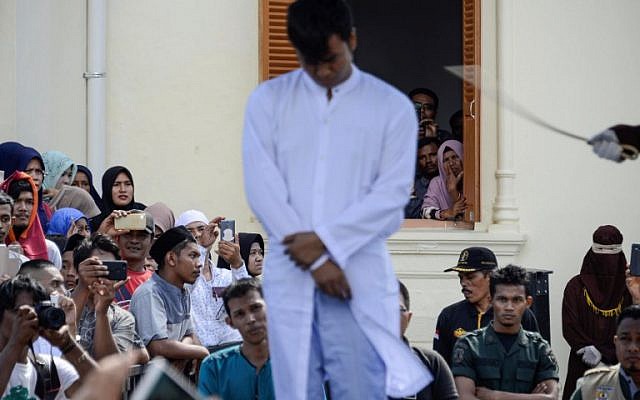 Onlookers watch as a member of Indonesia's Sharia police (R) whips a man (C) accused of having gay sex during a public caning ceremony outside a mosque in Banda Aceh, capital of Aceh province on July 13, 2018. (AFP/CHAIDEER MAHYUDDIN)